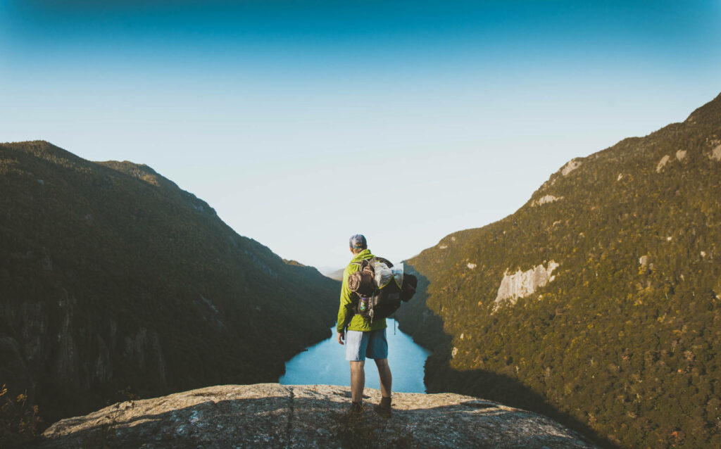 Solo travel tips. A solo traveller and backpacker stands looking out over a river in a valley.