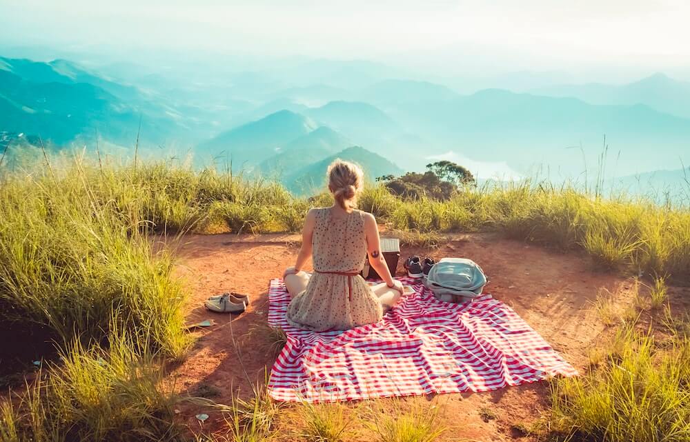 A solo female traveller sits on a picnic rug looking over a mountain range.
