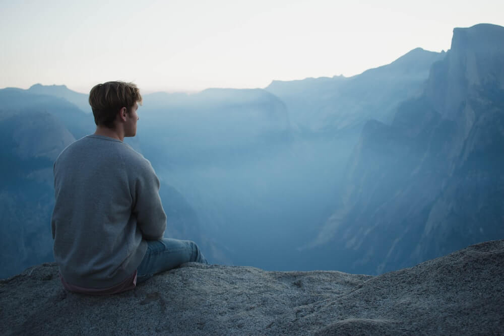 A male solo traveller sits on a cliff overlooking a mountainous valley.