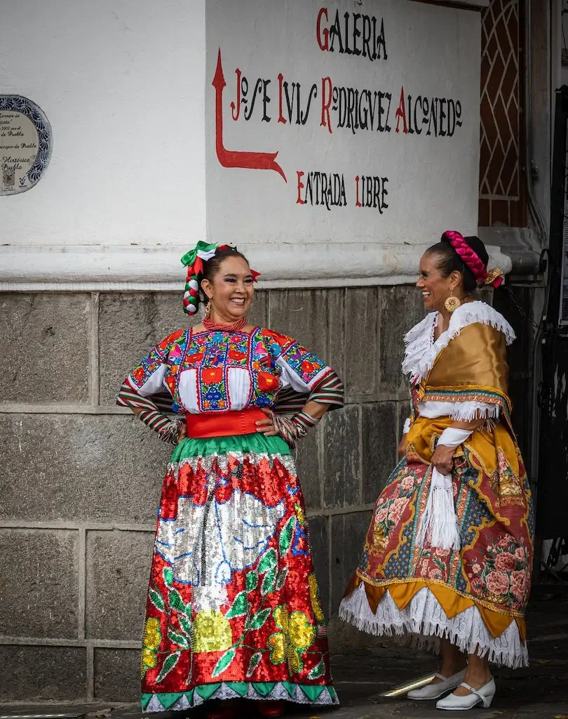 Two Mexican women dressed in huipil dresses on the street corner.