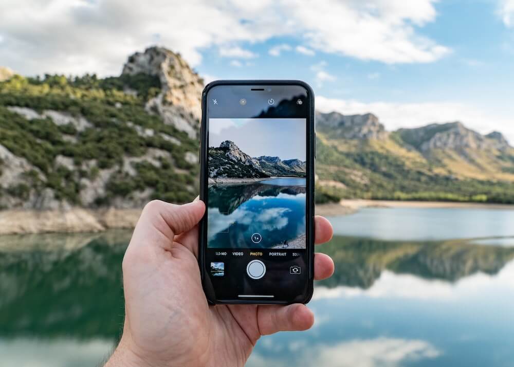 A person holds up their phone camera and takes a photo of a lake.