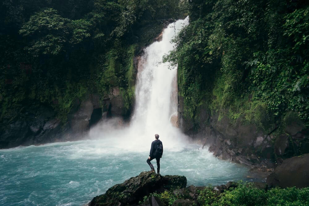 A solo male traveller stands at the base of a waterfall in a forest.