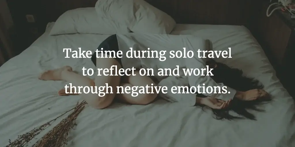 A lady curled up on a bed. The text reads, 'Take time during solo travel to reflect on and work through negative emotions.'