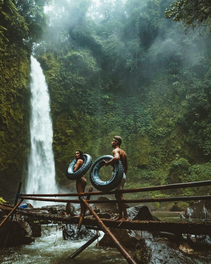 A couple travelling together stand at the bottom of a rainforest waterfall.