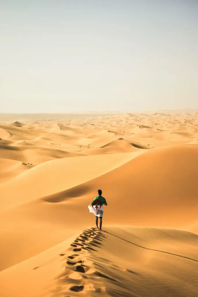 Solo male traveller wrapped in Algerian flag climbing sand dune in Sahara Desert, Algeria. 